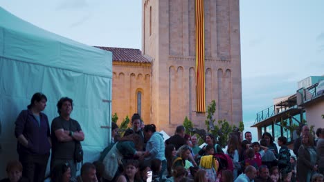 The-bell-tower-of-a-church-with-the-Senyera,-the-flag-of-Catalonia,-hanging-from-the-top-of-the-bell-tower,-falling-onto-a-square-full-of-people-on-Saint-George's-Day,-Sant-Jordi