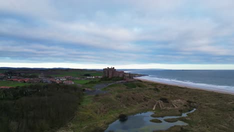 Pull-back-ascending-drone-footage-of-Bamburgh-Castle,-Northumberland-revealing-the-sandy-beach-and-coastline-beyond