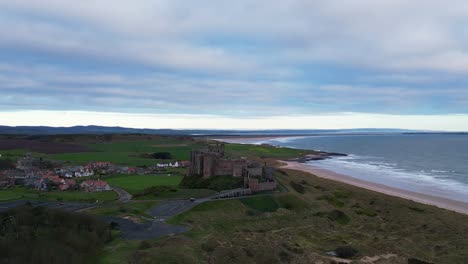 Descending-view-while-orbiting-anticlockwise-around-Bamburgh-Castle,-Northumberland-showing-the-stunning-location-beside-the-sea-and-Lindisfarne-Island-in-the-distance