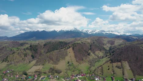 Gliding-Through-the-Mesmerizing-Vistas-of-Bucegi-Mountain-Range-A-Breathtaking-Drone-Journey-Featuring-Majestic-Snow-Clad-Summits,-Azure-Skies,-Fluffy-Clouds,-and-a-Serene-Mountain-Village,-Romania