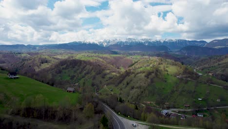Smooth-Aerial-View-Over-A-Mountain-Road-With-The-Majestic-Piatra-Craiului-Mountain-Range-In-The-Background,-Clear-Blue-Sky-With-Fluffy-White-Clouds,-Romania,-Transilvania,-Moieciu,-Rucar,-Bran