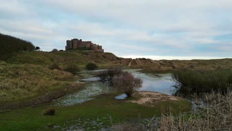 Camera-rises-from-a-swampland-in-sand-dunes-next-to-the-stunning-Bamburgh-Castle,-to-reveal-the-beauty-of-the-sea,-beach-and-surrounding-islands