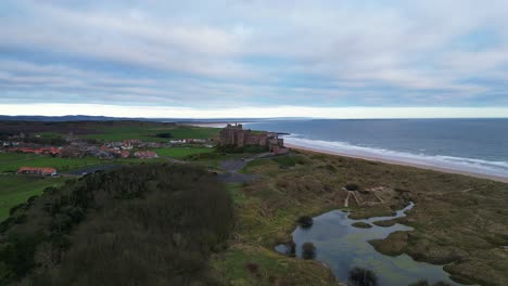 Clockwise-distant-rotate-aerial-footage-of-Bamburgh-Castle,-sand-dunes,-swamp-and-the-coastal-landscape-beyond