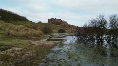 Close-to-the-ground-slow-flight-over-a-swamp-in-sand-dunes-at-the-foot-of-Bamburgh-Castle,-who's-reflection-can-be-seen-in-the-water
