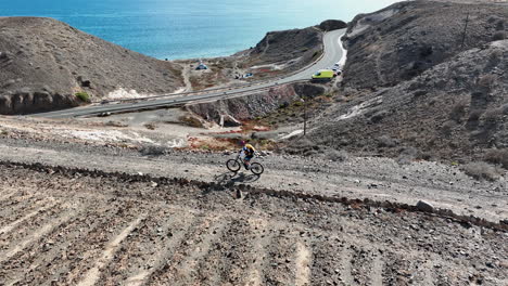 Lateral-aerial-shot-of-a-man-with-his-mountain-bike-in-a-desert-landscape-and-where-the-coast-and-the-sea-can-be-seen