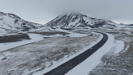 Driving-in-Winter-Landscape-of-Iceland,-Aerial-View-of-Red-Car-on-Wet-Road-by-Snow-Capped-Hills