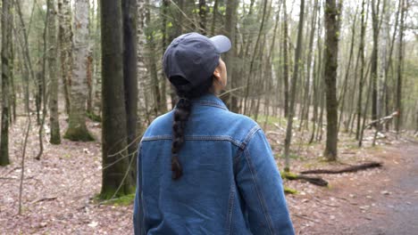 Young-diverse-woman-wearing-a-long-brunette-braid-jean-jacket-and-grey-baseball-cap-hat-walking-hiking-through-hike-trail-path-in-dense-forest-upstate-new-york-during-fall-spring-time-4k-slow-motion
