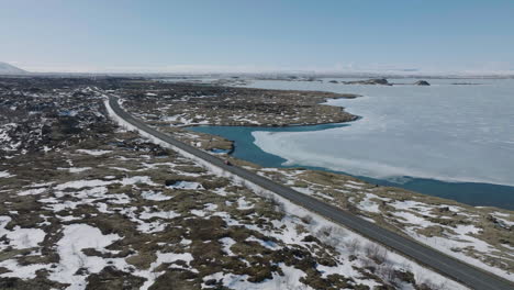 Aerial-View-of-Car-Moving-on-Road-by-Frozen-Lake-in-Landscape-of-Iceland-on-Sunny-Late-Winter-Day