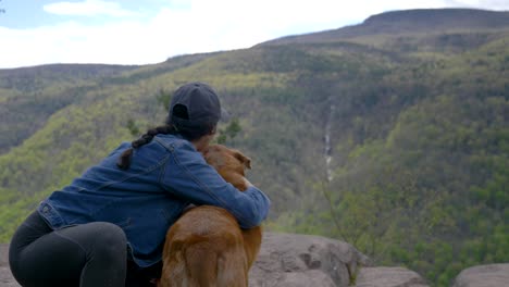 Young-diverse-woman-hiker-pets-her-brown-senior-dog-at-hiking-trail-viewpoint-rocky-cliff-overlooking-a-beautiful-picture-perfect-view-of-a-forest-mountainside-waterfall-4K-SLOW-MOTION-depth-of-field