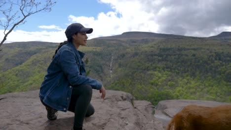 Pretty-young-woman-smiling-at-hiking-trail-viewpoint-overlooking-waterfall-smiles-at-bright-sunny-cliffside-view-waterfall-overlook-with-senior-happy-dog