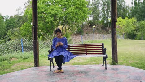 A-pretty-Asian-girl-wearing-glasses-on-a-bench-using-her-mobile-while-sitting-outdoors-on-a-park-bench,-Islamabad,-Pakistan