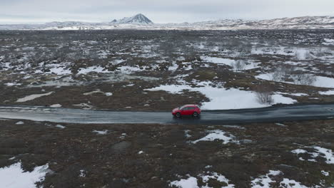 Aerial-View-of-Red-Car-Moving-on-Wet-Countryside-Road-in-Scenic-Landscape-of-Iceland,-Late-Winter-Season