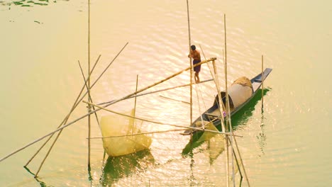Asiatischer-Mann-Steht-Bei-Sonnenuntergang-Auf-Einer-Bambusfischkonstruktion-In-Der-Nähe-Eines-Bootes,-Während-Die-Sonne-Untergeht