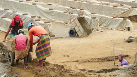 Asian-workers-collaborate,-moving-soil-filled-wheelbarrow-in-brick-factory