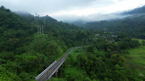 Aerial-drone-dolly-above-tropical-road-bridge-against-steep-green-mountain,-cloudy-day