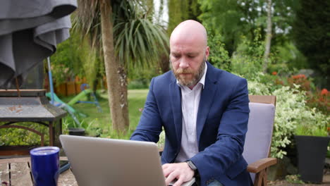 A-man-working-on-a-laptop-computer-in-garden-with-his-coffee-on-a-table-surrounded-by-trees,-plants-and-flowers-on-a-windy-day