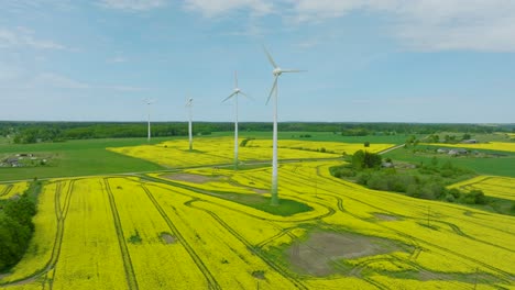 Aerial-establishing-view-of-wind-turbines-generating-renewable-energy-in-the-wind-farm,-blooming-yellow-rapeseed-fields,-countryside-landscape,-sunny-spring-day,-wide-drone-shot-moving-forward