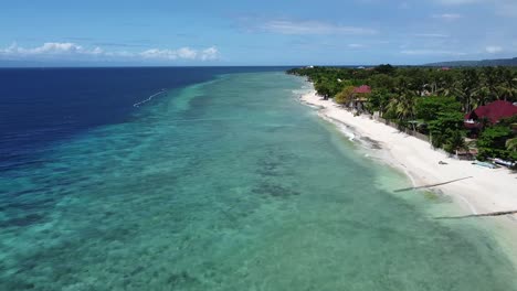 Mesmerizing-drone-footage-glides-along-Nacpan-Beach,-showcasing-vibrant-green-waters-with-coral,-sandy-shoreline,-palm-trees,-and-a-clear-blue-sky