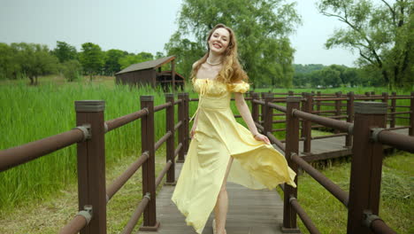 Lovely-woman-in-yellow-dress-walks-park-boardwalk,-seductive-looks-at-camera