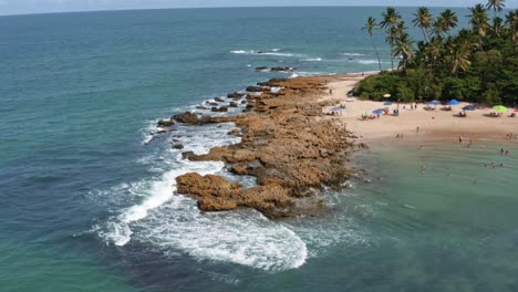 Trucking-right-aerial-drone-shot-of-small-waves-crashing-into-a-small-rock-peninsula-on-the-famous-Coqueirinhos-beach-in-Paraiba,-Brazil-with-tourists-and-locals-swimming-enjoying-the-water-and-sand