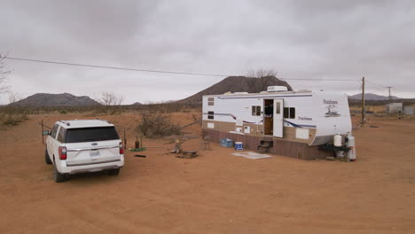 White-SUV-and-trailer-mobile-home-parked-on-dry-red-dirt-desert,-grey-storm-clouds-behind