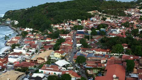 Dolly-in-tilt-down-aerial-drone-shot-of-the-famous-tourist-beach-town-of-Pipa,-Brazil-in-Rio-Grande-do-Norte-with-people-and-cars-passing-by-the-small-cobblestone-road-full-of-shops-and-food
