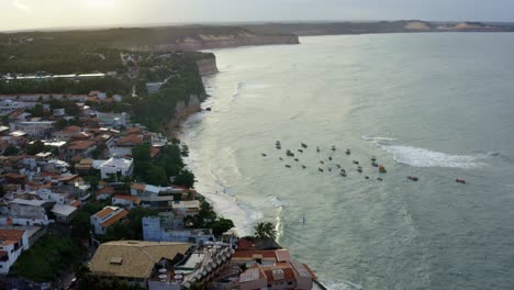Rotating-aerial-drone-shot-of-a-large-cluster-of-small-boats-in-the-water-of-the-famous-tropical-Pipa-beach-in-Rio-Grande-do-Norte,-Brazil-during-high-tide-surrounded-by-the-city-and-large-cliffs