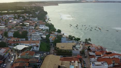 Tilting-down-aerial-drone-shot-of-a-large-cluster-of-small-boats-in-the-water-of-the-famous-tropical-Pipa-beach-in-Rio-Grande-do-Norte,-Brazil-during-high-tide-surrounded-by-the-city-and-large-cliffs