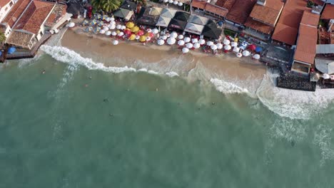 Rising-aerial-drone-birds-eye-shot-of-the-beautiful-tropical-famous-Pipa-beach-during-high-tide-with-tourists-playing-in-the-water-and-enjoying-the-shade-under-colorful-umbrellas-on-a-summer-evening