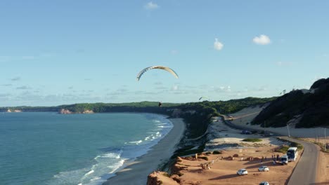 Aerial-drone-shot-of-a-paraglider-flying-over-the-stunning-tropical-northeastern-Brazil-coastline-near-Pipa-in-Rio-Grande-do-Norte-with-large-sand-dunes,-cliffs-over-the-ocean-and-green-foliage