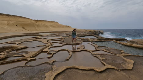 Mujer-Caminando-Por-La-Orilla-De-Una-Playa-Con-Charcos-De-Agua-En-La-Piedra-Erosionada