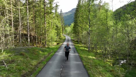 Man-jogging-towards-drone-camera-on-paved-road-through-lush-morning-forest