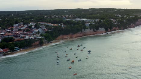 Rotating-aerial-drone-shot-of-a-cluster-of-small-fishing-boats-docked-on-the-tropical-Pipa-beach-during-high-tide-surrounded-by-large-cliffs,-homes-and-restaurants-in-Rio-Grande-do-Norte,-Brazil