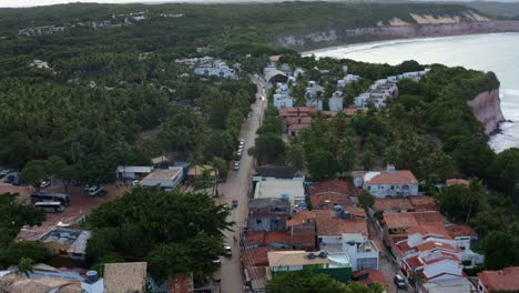 Dolly-in-aerial-drone-shot-of-the-main-street-leading-into-the-famous-tropical-tourist-town-of-Pipa,-Brazil-in-Rio-Grande-do-Norte-with-large-cliffs-above-the-calm-turquoise-ocean-and-green-foliage