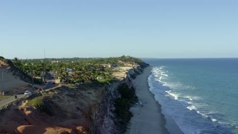 Truck-right-aerial-drone-shot-of-a-paraglider-flying-down-the-famous-tropical-Cacimbinhas-Cliffs-near-Pipa,-Brazil-in-Rio-Grande-do-Norte-with-large-sand-dunes,-green-foliage,-and-blue-ocean-water