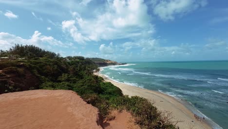 Tilt-up-handheld-shot-revealing-stunning-northeast-Brazil-coastline-in-Pipa,-Rio-Grande-do-Norte-on-top-of-a-large-orange-cliff-looking-down-at-the-famous-love-beach-with-golden-sand-and-small-waves