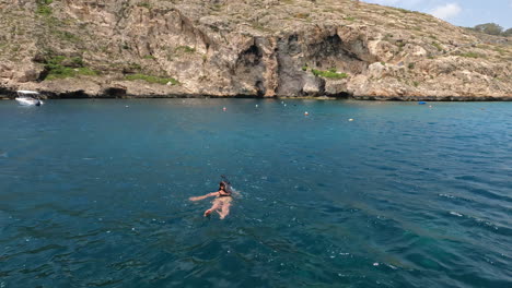 Woman-swimming-in-the-crystal-clear-water-next-to-a-coast-with-cliffs-and-some-boats