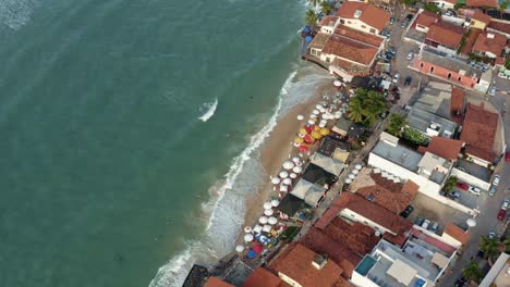 Rotating-aerial-drone-birds-eye-shot-of-the-beautiful-tropical-famous-Pipa-beach-during-high-tide-with-tourists-playing-in-the-water-and-enjoying-the-shade-under-colorful-umbrellas-on-a-summer-evening