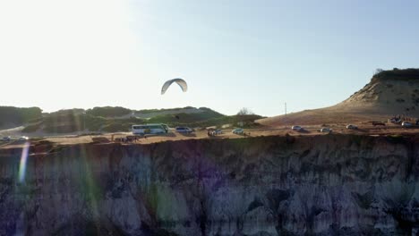 Rotating-aerial-drone-shot-following-a-paraglider-flying-along-a-cliff-with-large-sand-dunes-in-the-background-near-Pipa,-Brazil-in-Rio-Grande-do-Norte-on-a-warm-summer-day