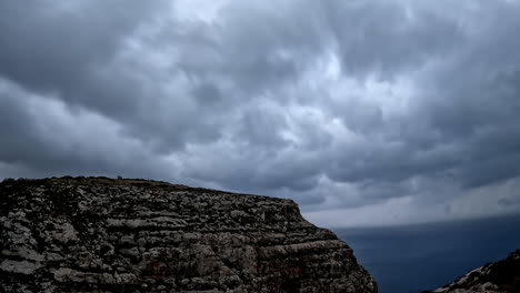 High-angle-shot-over-beautiful-blue-oceanic-beach-from-rocky-cliff-with-dark-rain-clouds-passing-by-in-timelapse