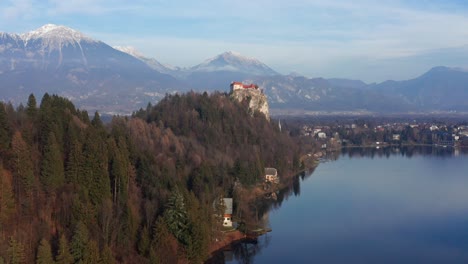 Aerial-Flying-Towards-Lake-Bled-Castle-With-Snow-Capped-Karawanks-Mountain-Range-In-Background