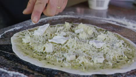 Chef-Agregando-Queso-Mozzarella-Y-Coberturas-A-La-Pizza-Casera-En-Una-Cocina-Rústica-Al-Aire-Libre