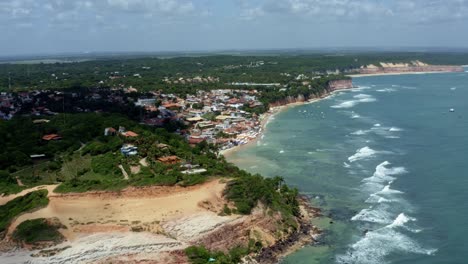 Right-trucking-extreme-wide-aerial-drone-landscape-shot-of-the-famous-tropical-tourist-beach-town-of-Pipa,-Brazil-in-Rio-Grande-do-Norte-with-small-waves,-cliffs,-golden-sand,-and-green-foliage