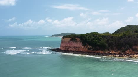Dolly-out-aerial-drone-shot-of-a-large-tropical-cliff-over-the-ocean-revealing-small-dolphin-tour-boats-anchored-with-tourists-swimming-near-the-famous-Madeiro-beach-in-Pipa-Brazil-Rio-Grande-do-Norte