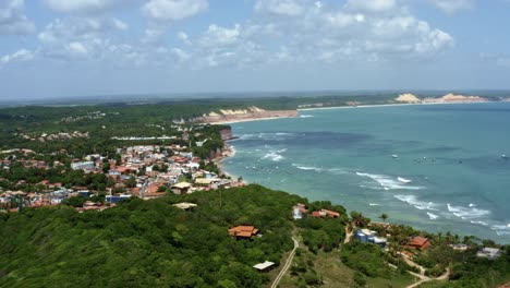Left-trucking-extreme-wide-aerial-drone-landscape-shot-of-the-famous-tropical-tourist-beach-town-of-Pipa,-Brazil-in-Rio-Grande-do-Norte-with-small-waves,-cliffs,-golden-sand,-and-green-foliage