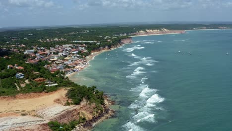 Left-trucking-extreme-wide-aerial-drone-landscape-shot-of-the-famous-tropical-tourist-beach-town-of-Pipa,-Brazil-in-Rio-Grande-do-Norte-with-small-waves,-cliffs,-golden-sand,-and-green-foliage