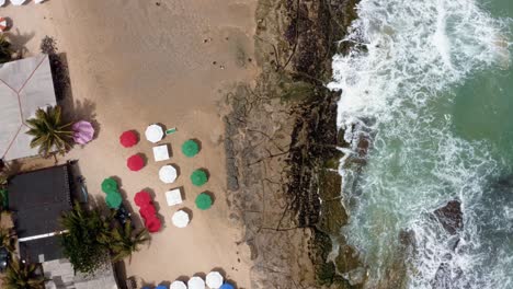 Aerial-drone-birds-eye-top-view-of-small-waves-crashing-into-a-cliff-of-rocks-from-the-tropical-popular-Tibau-do-Sul-beach-near-Pipa,-Brazil-in-Rio-Grande-do-Norte-with-colorful-umbrellas-for-tourists