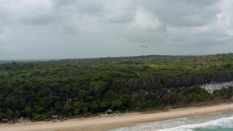Left-trucking-aerial-drone-shot-of-a-lone-paragliding-flying-over-the-famous-tropical-Madeiro-beach-near-Pipa,-Brazil-in-Rio-Grande-do-Norte-with-large-looming-green-cliffs,-golden-sand-and-blue-water