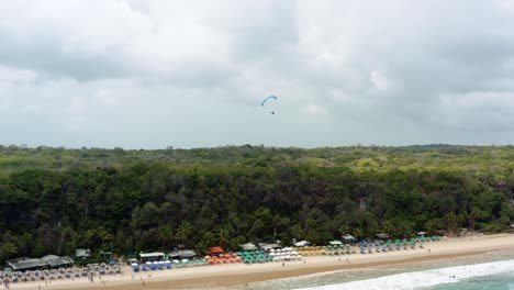 Left-trucking-aerial-drone-shot-of-a-lone-paragliding-flying-over-the-famous-tropical-Madeiro-beach-near-Pipa,-Brazil-in-Rio-Grande-do-Norte-with-large-looming-green-cliffs,-golden-sand-and-blue-water