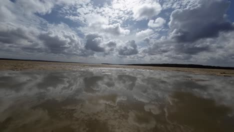 Dolly-in-landscape-worms-eye-view-shot-of-a-tropical-wet-sand-bar-during-low-tide-in-the-Guaraíras-Lagoon-of-Tibau-do-Sul,-Brazil-in-Rio-Grande-do-Norte-during-a-sunny-summer-cloudy-day-near-Pipa